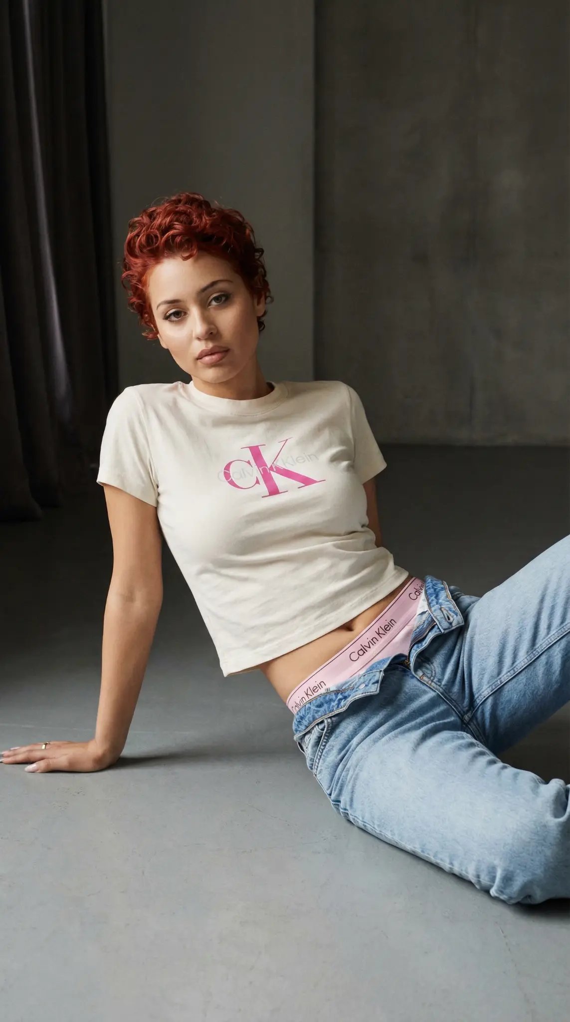 Calvin Klein underwear campaign with model in pink CK logo tee and visible waistband lounging on gray studio floor